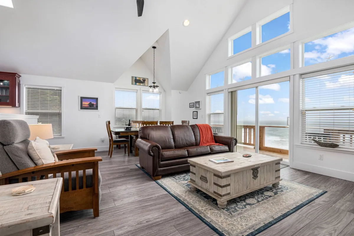 The image showcases a bright, inviting open living space with plush leather sofas and a wooden coffee table, all positioned to take in sweeping ocean views through expansive windows. The dining area is nearby, offering a picturesque spot to savor meals while gazing at the Pacific. The overall ambiance is cozy and welcoming, ideal for relaxing or socializing after a day at the beach.