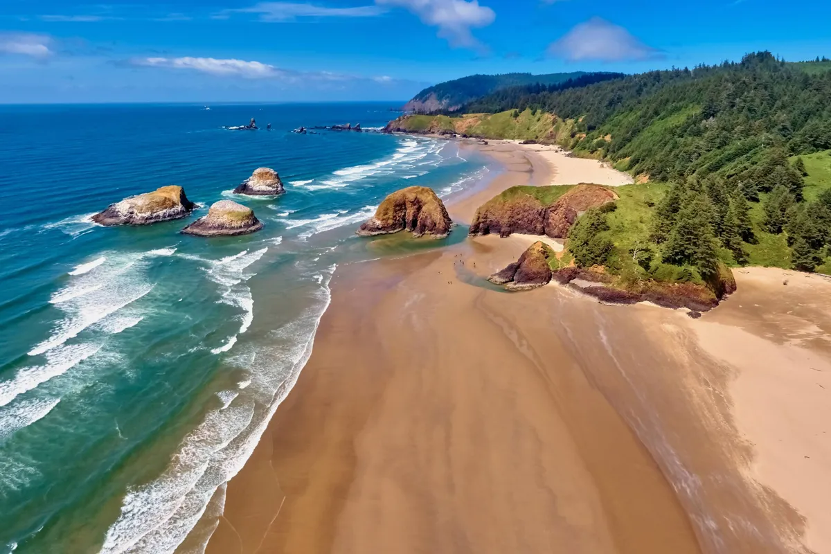 A wide sandy beach framed by rock formations and lush coastal greenery, with gentle waves rolling in under a soft, glowing sky. The environment looks perfect for family activities such as swimming, sunbathing, building sandcastles, and beachcombing. The lush greenery adds a natural, inviting touch to the beach scene.