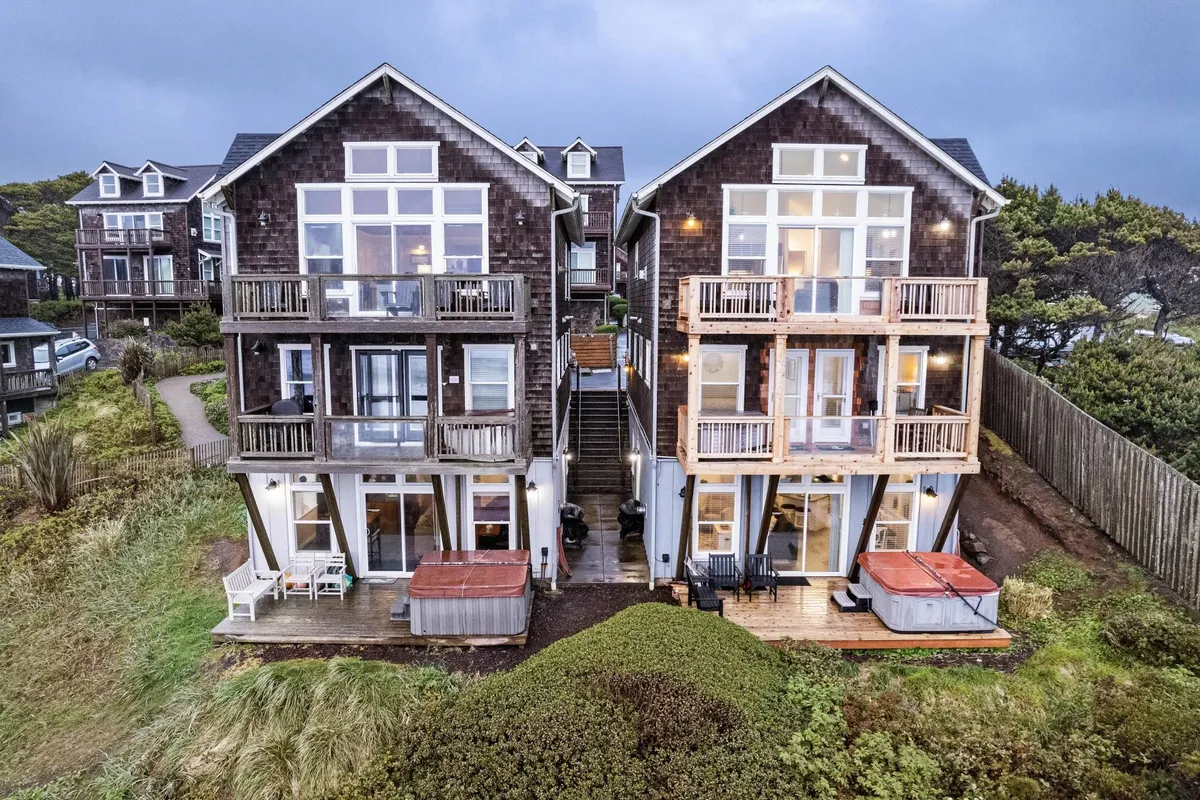 This exterior shot shows two adjacent houses, each with large windows, hot tubs, and spacious wooden decks. The ocean is visible beyond, and the decks are set up for gatherings or quiet enjoyment of the sunset. The scene conveys a lifestyle of relaxation, with options for both private and social outdoor enjoyment.