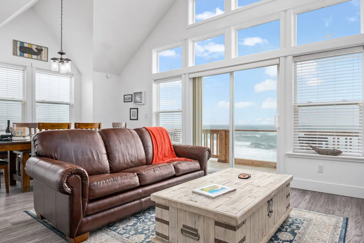 This interior image showcases a leather sofa and a wooden coffee table positioned before large windows with unobstructed ocean views. The relaxed and welcoming space invites moments of comfort, whether sipping coffee, reading, or simply gazing at the rolling waves.