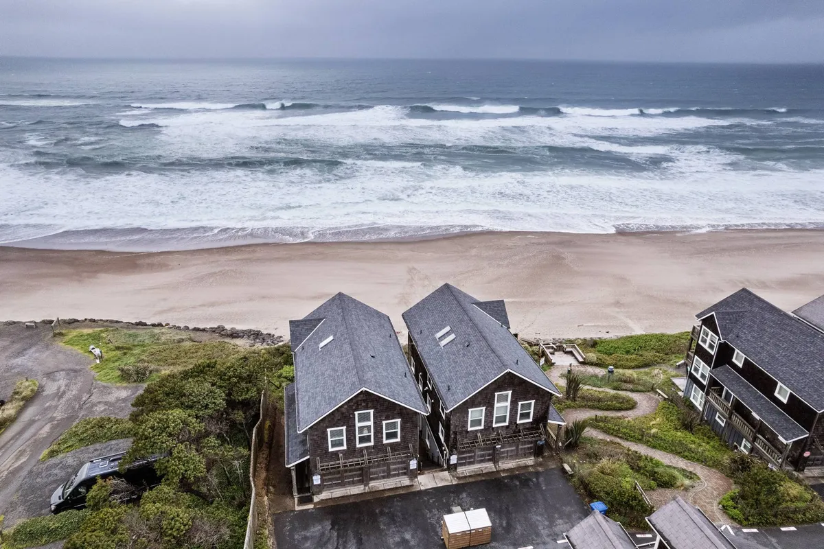 An exterior shot featuring two neighboring beach houses facing the ocean, with sandy beach and waves visible in the background. The setting conveys a tranquil, coastal vibe and hints at the proximity of adjacent accommodations for larger groups.