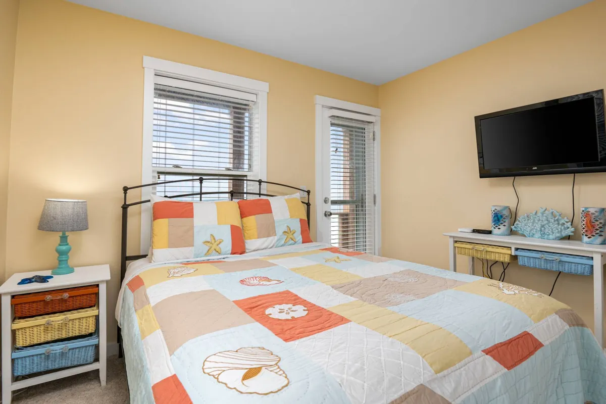 A cozy bedroom with a bed dressed in a colorful quilt, blue lamp, wall-mounted television, and a decorative side table. Natural light streams in, creating a warm and restful environment ideal for winding down with a favorite show or enjoying quiet moments.