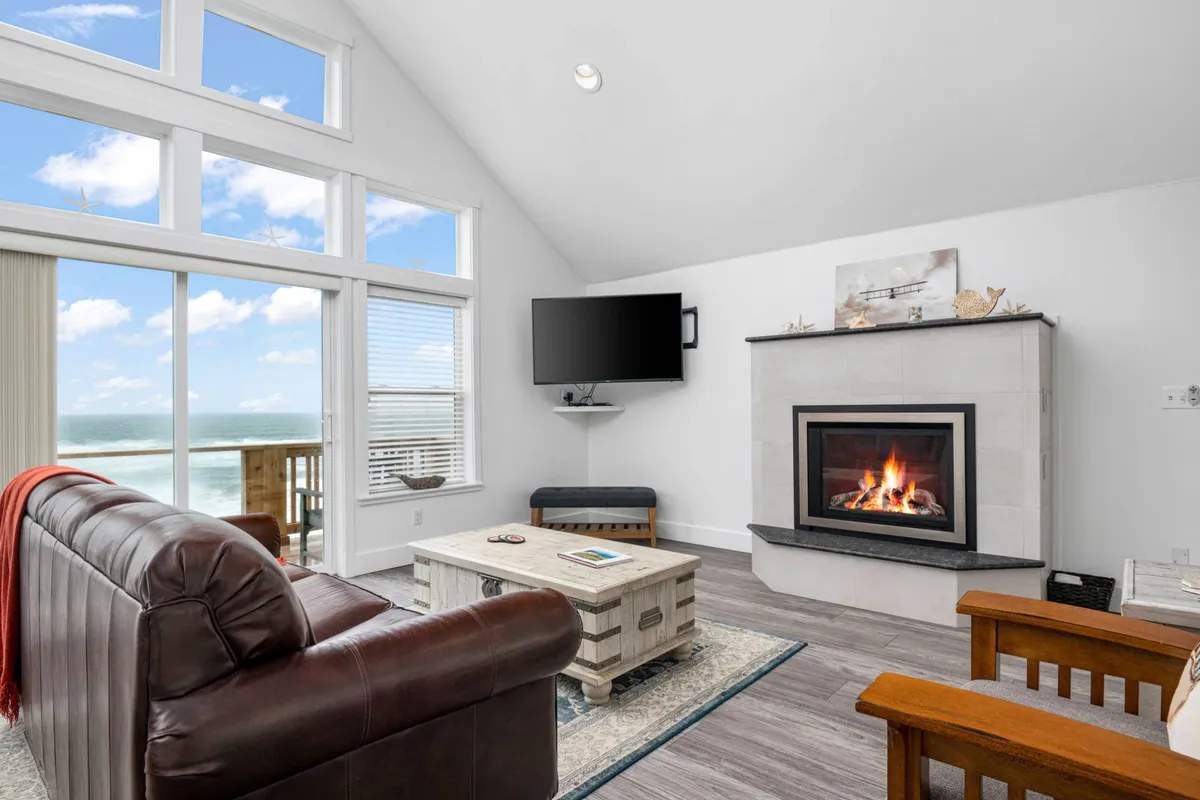 A bright and spacious living room with a leather sofa and coffee table arranged in front of a modern gas-log fireplace. Large windows dominate one wall, flooding the space with natural light and offering sweeping ocean views. The space exudes comfort and warmth, enhanced by tasteful décor and a serene ambiance perfect for relaxing or socializing.