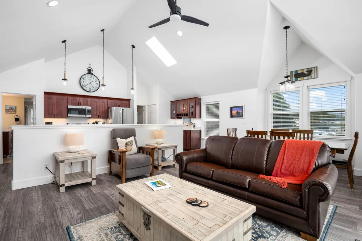 A welcoming open-concept space featuring a leather sofa, wooden coffee table, visible kitchen area, and a dining table ready for gatherings. The layout encourages social interaction, and the space is bathed in natural light from unseen windows, reinforcing a sense of connection and warmth.