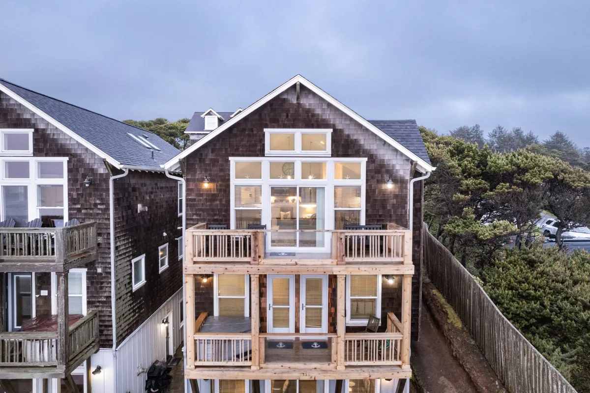 This exterior view centers on the home’s large front windows and wooden balconies, with the shingled exterior lending a classic coastal vibe. The balconies are positioned for sunset views and include outdoor seating, hinting at intimate conversations or solo moments with a coffee in the fresh air.