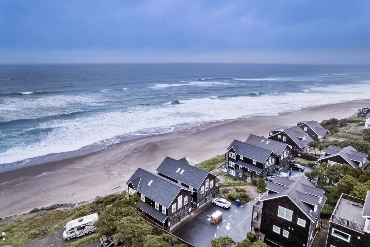 A scenic vista of beachfront houses nestled along the sand dunes, with waves crashing and a tranquil, open beach stretching into the distance. The scene evokes the expansive, natural beauty of the coast and hints at the walkable access from the home to the shoreline.