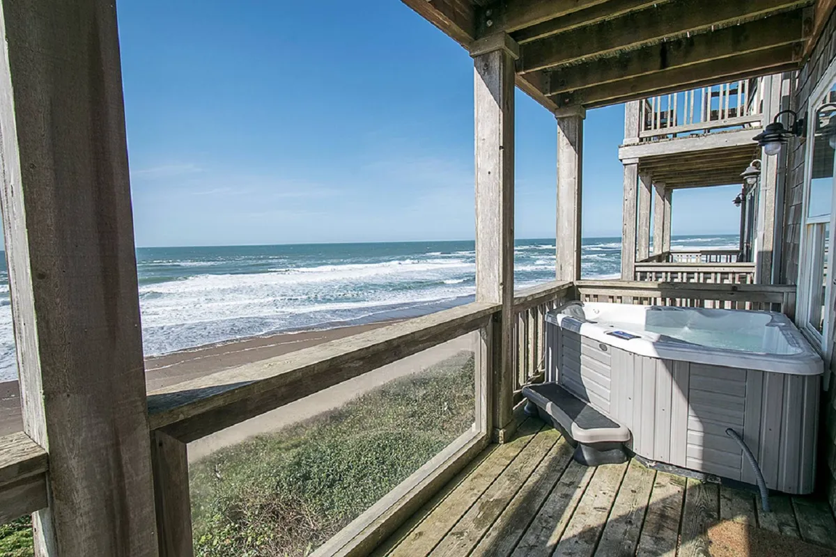 This image highlights a private hot tub set on a wooden deck, with sweeping views of the ocean visible beyond the railing. The mood is serene, with the hot tub invitingly steaming and the expansive water stretching toward the horizon, suggesting sunset or twilight. Outdoor seating is subtly visible, reinforcing the idea of relaxed gatherings or solo retreats.