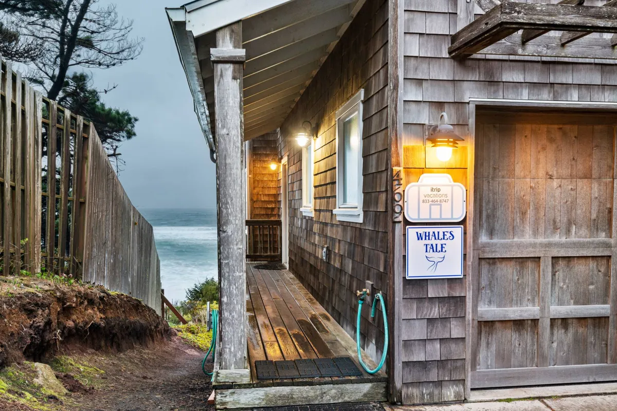 The exterior of the Whale’s Tale property, featuring wooden siding and a sign with 'WHALES TALE.' The porch is warmly lit, and the ocean is visible in the background, reinforcing proximity to the beach. The scene suggests a relaxing spot to enjoy the view or set off for seaside adventures.