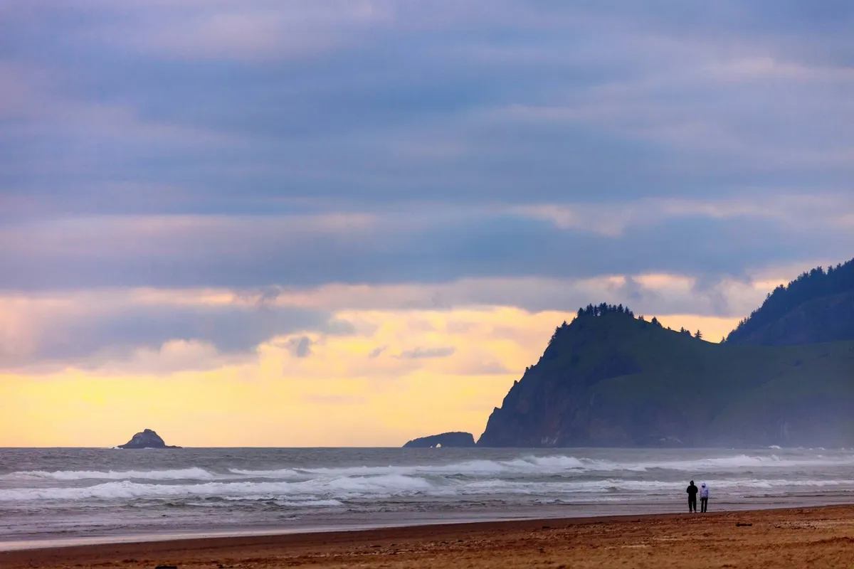 A serene beachscape featuring gentle ocean waves, a dramatic cliff silhouette, and two friends walking together along the shoreline as the sun sets. The mood is peaceful and intimate, evoking a sense of shared adventure and relaxation.